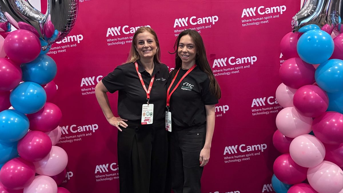 Samantha and Annie standing in front of a bright pink backdrop that says 'A11y Camp' with pink and blue balloons either side.  Samantha and Annie are both wearing black shirts and smiling.