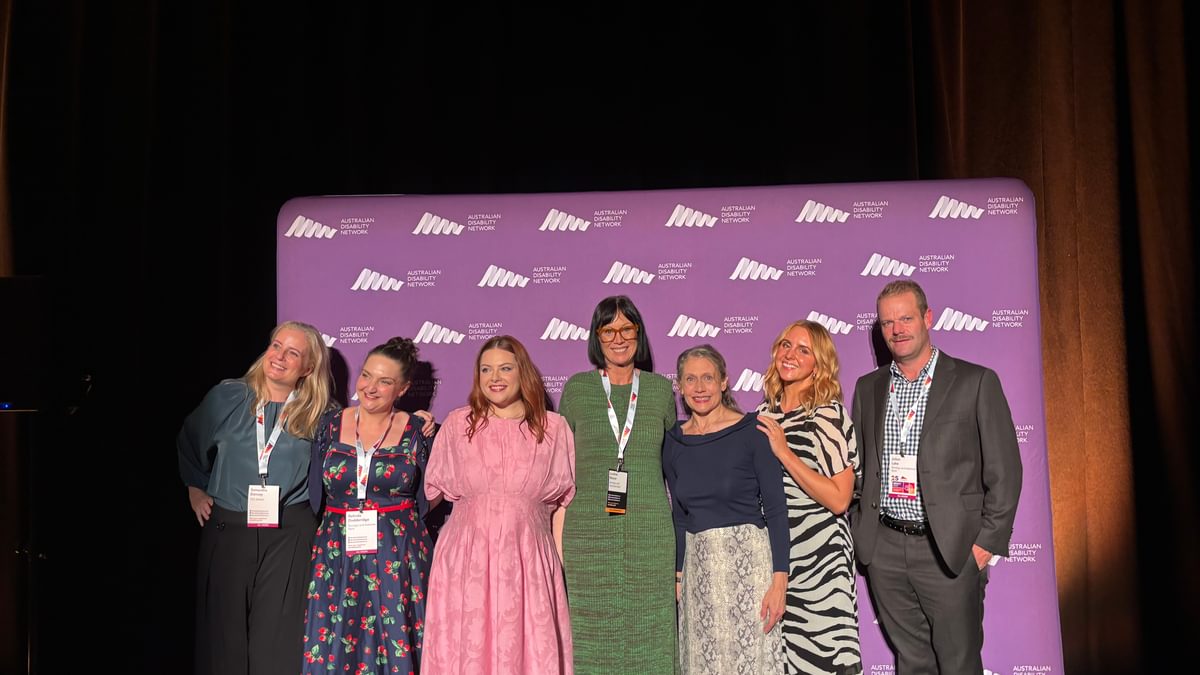 A group of seven people standing in front of a purple background with the logo for Australian Network on Disability. They are smiling and looking at the camera.