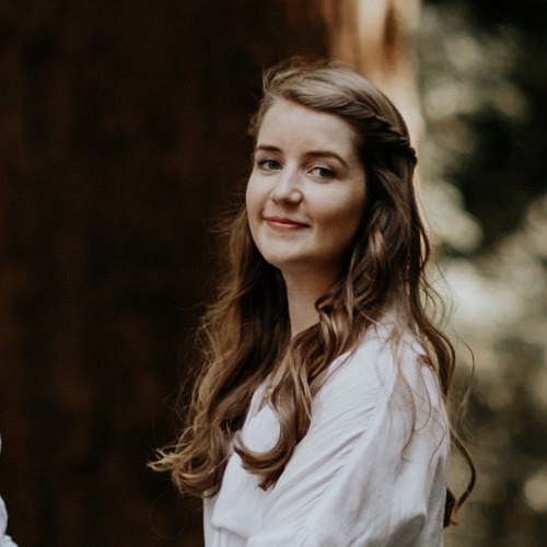 Karla has long brown hair and is wearing white, smiling in the outdoors