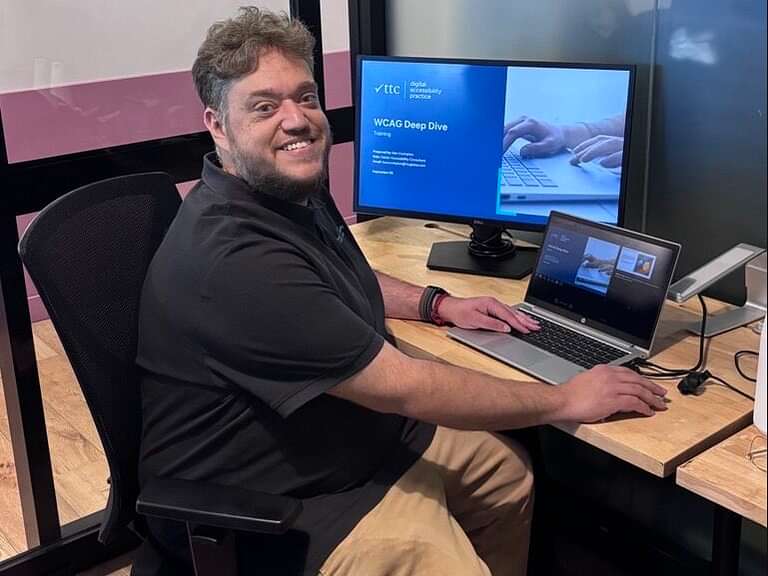 Person sitting at a desk smiling looking at the camera. He is sitting in front of a laptop monitor that says WCAG Deep Dive on the screen