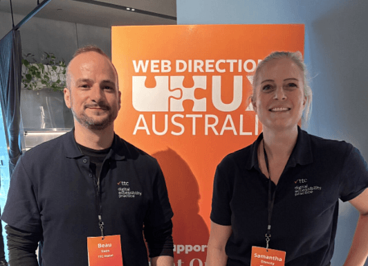 Beau and Samantha standing in front of the Web Directions UX Australia Banner Smiling