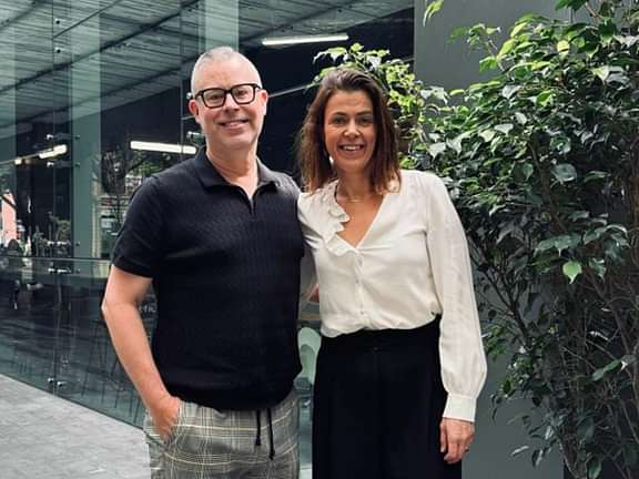 A man and woman standing next to each other and smiling. The man has grey hair, black glasses and is wearing a black shirt. The woman has brown hair, a white shirt and black skirt.