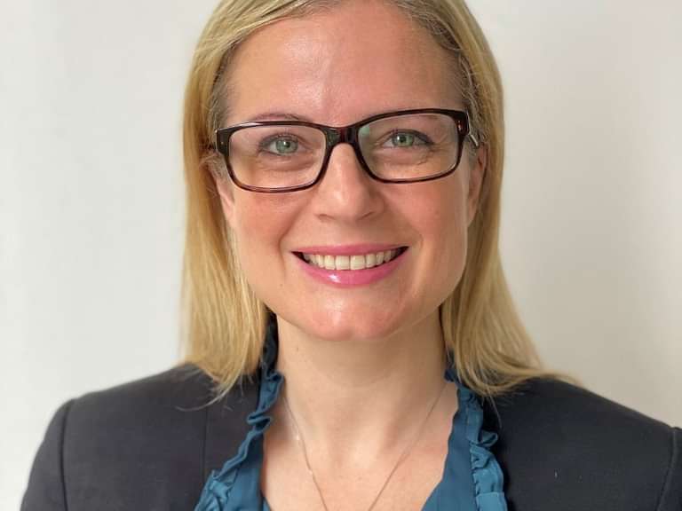 Sam is a caucasian female with shoulder length blonde hair and grey eyes. Wearing glasses and a green shirt with a black jacket